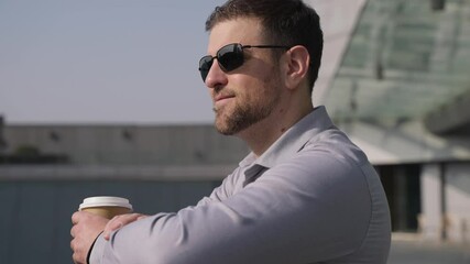 Wall Mural - Successful confident caucasian male businessman wearing sunglasses standing on the balcony looking forward holding coffee cup enjoy the sunshine afternoon