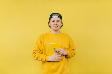 Wall Mural - Positive student guy with notebook and pen in hands isolated on yellow background, looking at camera with smile on face.