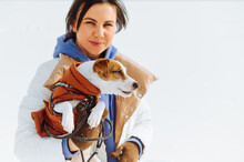 Portrait Of A Beautiful Woman With A Dog In A Jacket In His Hands On A White Snowy Background During A Winter Walk.