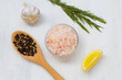 Pink Himalayan salt on the glass bowl, rosemary, piece of lemon, fresh garlic and mix of peppers in a wooden spoon on the white background. Top view copy space. Set of various spices for cooking.