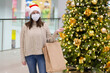 Woman wearing a facemask in a santa claus hat, sweater in a shopping center near the christmas tree. Concept: holiday shopping during the coronavirus epidemic a woman wearing a medical mask 