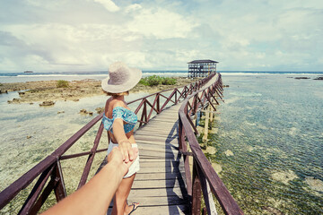 Wall Mural - Follow me. Travel concept. Vacation on tropical island. Back of young woman in hat holding boyfriend's hand enjoying sea view from wooden bridge terrace, Cloud 9 Surfing Spot, Siargao Philippines.