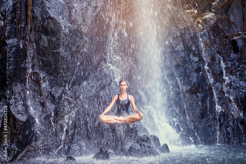 Wellness spa, vacation and yoga meditation concept. Pretty young woman in swimsuit sitting in lotus position on the rock under tropical waterfall.