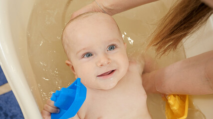 Wall Mural - Mother supporting and holding her little baby son while washing him in small bath. Concept of children hygiene, healthcare and parenting.