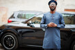 © AS Photo Family - African guy in blue authentic costume, eyeglasses and beret. Fashionable nigerian man near black suv car.