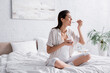 © LIGHTFIELD STUDIOS - cheerful pregnant woman holding glass jar and eating yogurt while sitting on bed.