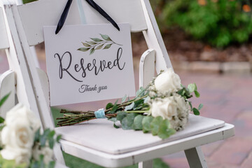 Closeup shot of a reserved wooden sign hanging on a white chair at a wedding ceremony Reserved seating for the family at an outdoor wedding ceremony A bouquet of white roses lies on the chair