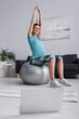 © LIGHTFIELD STUDIOS - positive pregnant woman with raised hands exercising on fitness ball and looking at laptop in living room.