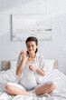 © LIGHTFIELD STUDIOS - pleased young pregnant woman holding glass jar with yogurt while sitting on bed.