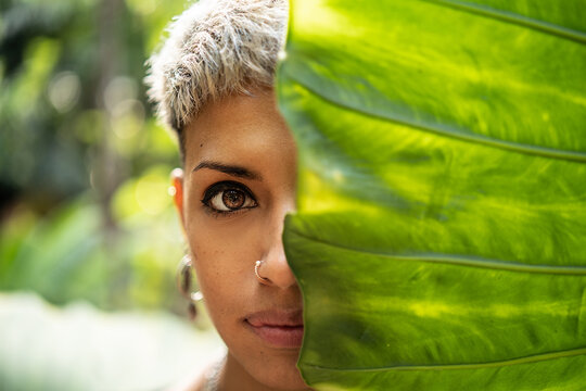 Portrait of beauty young woman looking trough green tropical leaf. Young woman's face surrounded by leaves. Fashionable girl with short hair and nose ring.