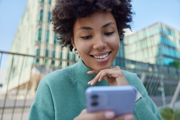 Wall Mural - outdoor shot of happy curly haired woman checks newsfeed on smartphone smiles gladfully dressed in c