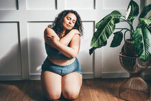 Curly Haired Overweight Young Woman In Blue Top And Shorts With Satisfaction On Face Accepts Curvy Body Shape In Stylish Bedroom.