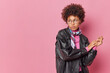 © WHstudio Leushin N - Serious pensive curly haired young woman wears leahter jacket kerchief around neck keeps hands together focused away isolated over pink background with copy space for your promotional content
