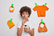 Indecisive thoughtful curly haired woman concentrated above holds chin thinks about something holds mobile phone considers selling clothes and unnecessary items isolated over white background