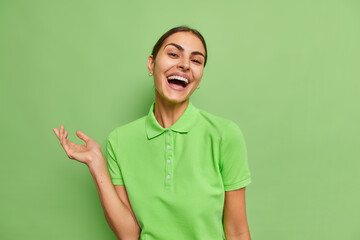 Wall Mural - Waist up shot of happy brunette woman dressed in casual t shirt smiles sincerely keeps arm raised laughs at funny anecdote wears casual t shirt isolated over green background. Happiness concept