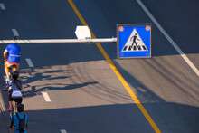 High Angle View And Focus At Overhead Electric Pedestrian Crossing Sign Over With Blurred Background Of Cyclist Group Riding On The Road At Morning Time