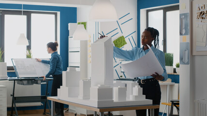 Poster - Engineer holding blueprints plan and analyzing building model for architecture work. Woman architect working with tools to design construction layout and structure for urban project.