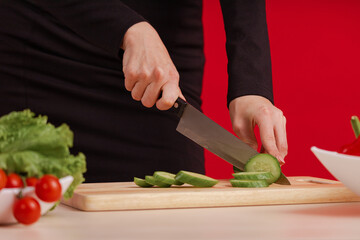 Female hands cut fresh cucumber on a wooden board on the table with a large cook knife on a red background