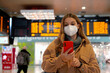 Traveler woman wearing FFP2 KN95 face mask at train station. Female commuter checking her smartphone with behind timetables of departures arrivals.