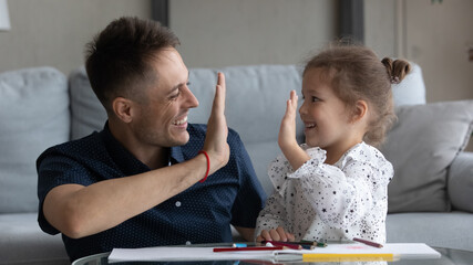 Happy excited dad and daughter girl giving high five over album with drawings, colorful pencils. Father showing praise, support for good homework result, playing creative learning games with child