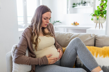 Wall Mural - Pregnant woman suffering during her pregnancy, with back pain and headaches. Pregnant woman sitting on the sofa holding her belly with worried face expression.