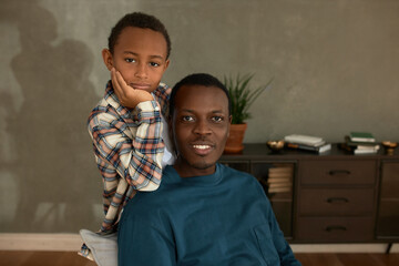 Wall Mural - Father and son relationships. Portrait of handsome dark-skinned boy in plaid shirt leaning on dad's shoulder, both having happy, calm face expression, feeling bonding, isolated on lounge interior