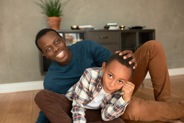 Wall Mural - Indoor image of father and son resting after lunch on floor in living-room, black ethnicity boy making funny faces while his dad holding his hand on head, showing his attitude and love