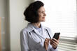 © fizkes - Happy pensive middle aged doctor woman using online app, medical service on smartphone, consulting internet, making appointment with patients, looking out of window, smiling, thinking