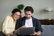 © fizkes - Adult son teaching mature mom to use online virtual app, banking service on laptop. Senior mother and grown child sitting on couch together, holding computer, browsing internet, talking