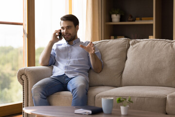 Canvas Print - Happy millennial business man speaking on cellphone, making telephone call from home, giving consultation to client, sitting, relaxing on comfortable couch. Distance work, communication concept