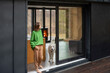 © rh2010 - Young woman with her dog at modern house on nature. Girl in green sweater standing near the window and look outside. Concept of comfort and happy modern life