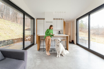 Young woman with her dog at modern house with panoramic windows. Concept of comfort and happy life at modern living spaces. Wide interior view