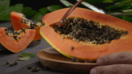 Wall Mural - Half papaya fruit with seeds on a vintage table with leaves of tropical plants. A hand with a wooden spoon cleans the papaya fruit from the seeds. Horizontal slow movement of the camera from left to r