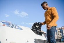 Urban, Electric Vehicle, Eco Concept. Young Black Skinned Man, Waiting For His Electric Car Charging