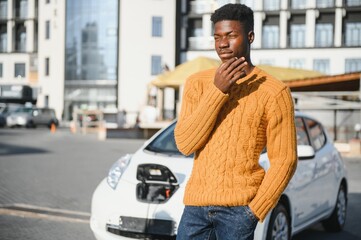 Wall Mural - Urban, electric vehicle, eco concept. Young black skinned man, waiting for his electric car charging