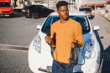 Wall Mural - african man holding charge cable in on hand standing near luxury electric car.