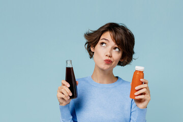 Young minded woman 20s wear casual sweater hold soda cola water vegetable smoothie choose between junk food and healthy isolated on plain pastel light blue background. People lifestyle food concept.