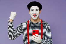 Charismatic Fun Young Mime Man With White Face Mask Wears Striped Shirt Beret Hold In Hand Use Mobile Cell Phone Doing Winner Gesture Isolated On Plain Pastel Light Violet Background Studio Portrait.