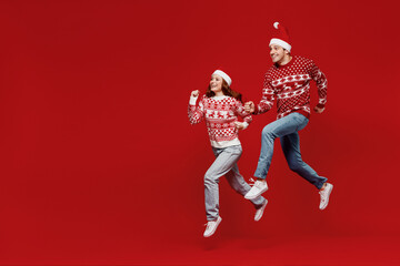 Wall Mural - Full body young couple friends two man woman 20s in sweater hat jump high run hurry up isolated on plain red background studio portrait. Happy New Year 2022 celebration merry ho x-mas holiday concept.