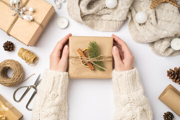 Wall Mural - First person top view photo of hands in sweater holding craft paper giftbox with twine bow pine twig cinnamon stick dried lemon slice scarf balls and tools for handicraft on isolated white background