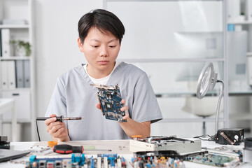 Poster - Young serious Asian woman with soldering iron looking at hardware while repairing it in workshop