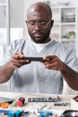 Poster - Young repairman of African ethnicity holding smartphone over hardware while sitting by workplace