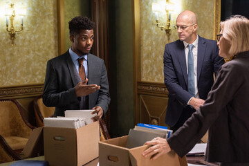 Poster - Young lawyer explaining something to one of mature colleagues while taking folder with papers out of box