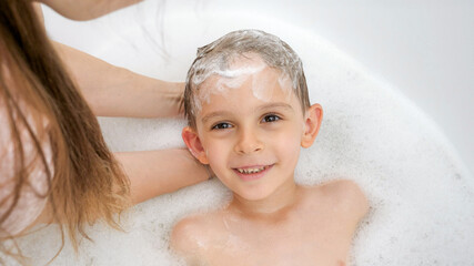 Wall Mural - Portrait of smiling little boy looking at mother while she washes his hair with shampoo in bathroom. Concept of hygine, children development and fun at home