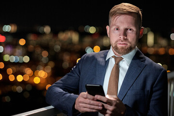 Poster - Young serious businessman with smartphone standing on balcony against night lights and looking at camera