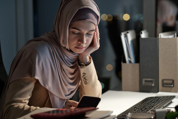 Poster - Young contemporary businesswoman scrolling in smartphone while sitting by workplace in dark office at night