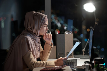 Wall Mural - Side view of young businesswoman in hijab looking through papers by workplace in office at night time