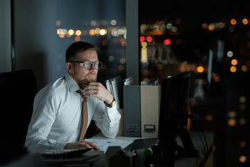 Poster - Serious broker or economist looking at computer screen while sitting by desk in dark office at night