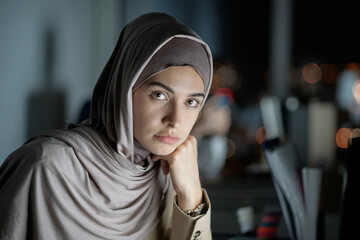 Poster - Young serious Muslim businesswoman in hijab looking at camera while sitting in office late at night