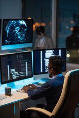 young african developer sitting in armchair by desk and typing while looking at coded data on comput